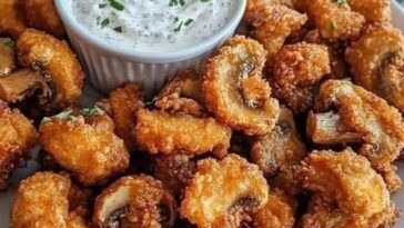 A plate of golden, crispy fried mushrooms served with a side of creamy cool ranch dip and garnished with fresh herbs.