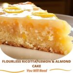 A slice of flourless ricotta, lemon, and almond cake on a white plate, topped with powdered sugar and a lemon zest garnish.