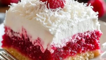 A slice of Raspberry Zinger Poke Cake, with vibrant raspberry filling soaked into the cake, topped with fluffy white frosting and fresh raspberries, served on a decorative plate.