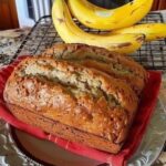 Slice of soft and moist banana bread on a wooden cutting board with a few ripe bananas beside it