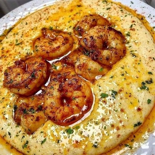 A bowl of creamy grits topped with Cajun-seasoned shrimp, garnished with chopped green onions and a sprinkle of paprika, served with a drizzle of butter sauce.
