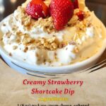Bowl of creamy strawberry shortcake dip topped with crushed golden cookies and fresh strawberry slices, surrounded by dippers like vanilla wafers and graham crackers.