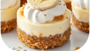 Mini banana pudding cheesecakes topped with whipped cream and banana slices, served in individual portions on a dessert tray.