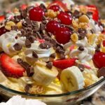 "A bowl of banana split fluff salad, with slices of banana, whipped cream, maraschino cherries, and colorful sprinkles, served with a spoon."