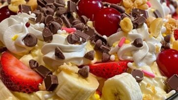 "A bowl of banana split fluff salad, with slices of banana, whipped cream, maraschino cherries, and colorful sprinkles, served with a spoon."