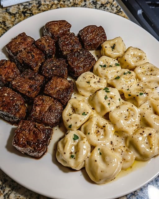 Garlic butter steak bites served with cheese-filled tortellini in a creamy Alfredo sauce, garnished with herbs.