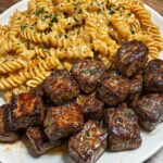 A plate of garlic butter steak bites served alongside twisted pasta coated in creamy spicy Cajun Alfredo sauce, garnished with parsley.