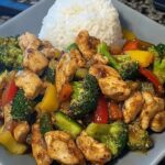 A colorful garlic chicken and vegetable stir-fry served over a bed of creamy rice on a white plate.