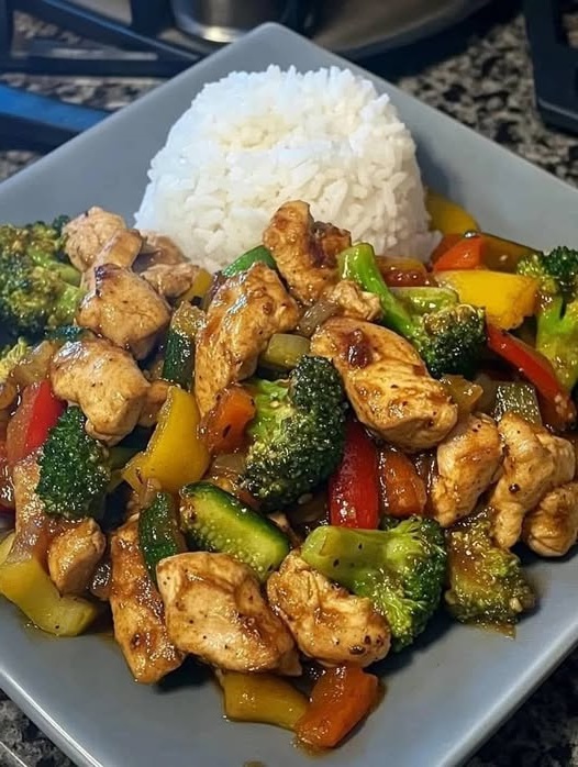 A colorful garlic chicken and vegetable stir-fry served over a bed of creamy rice on a white plate.