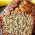 A moist slice of banana cake topped with creamy frosting and garnished with banana slices and a sprinkle of chopped nuts.